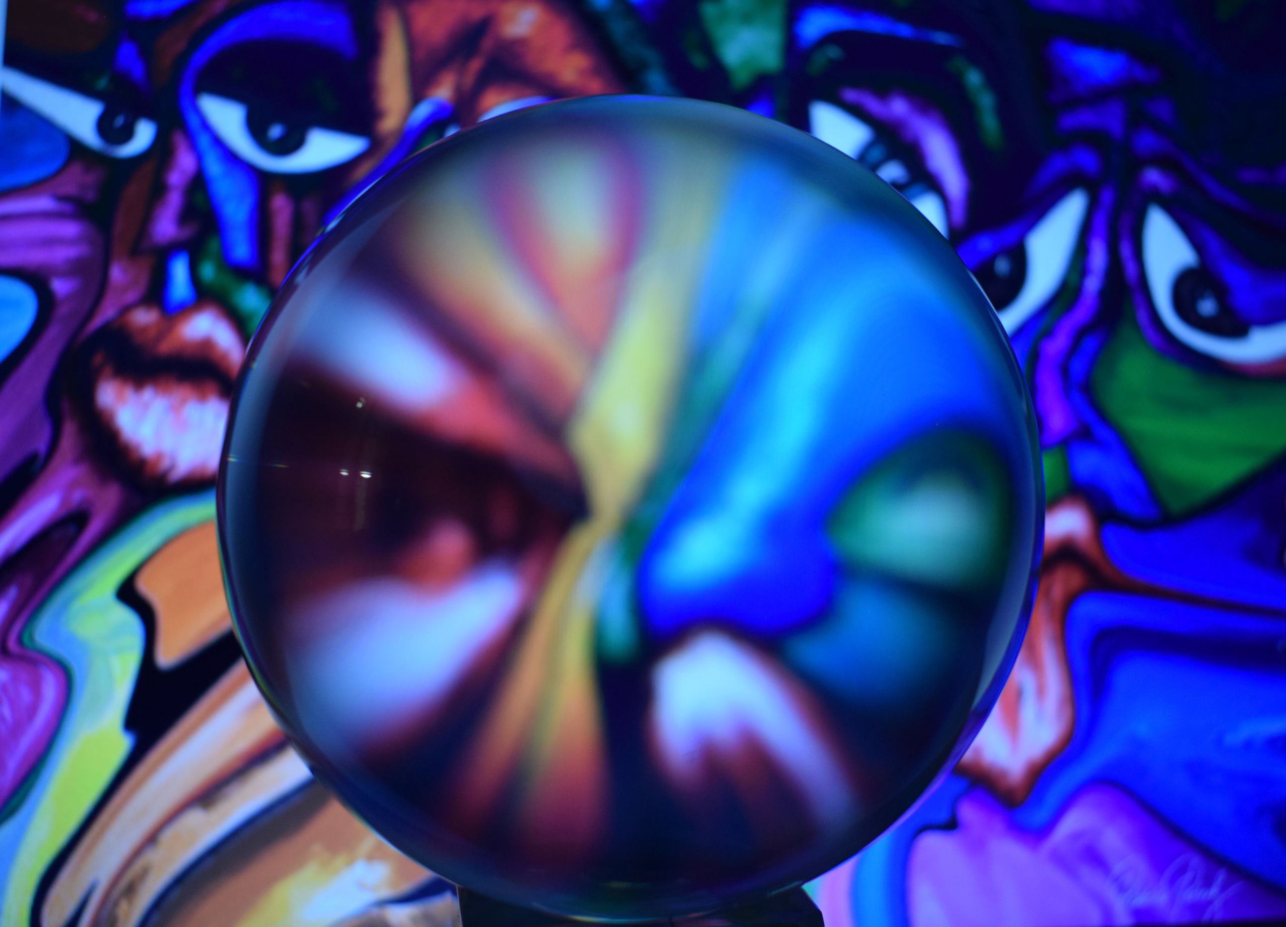 A glass sphere in front of abstract art of faces. Within the sphere the image and colours blend together.